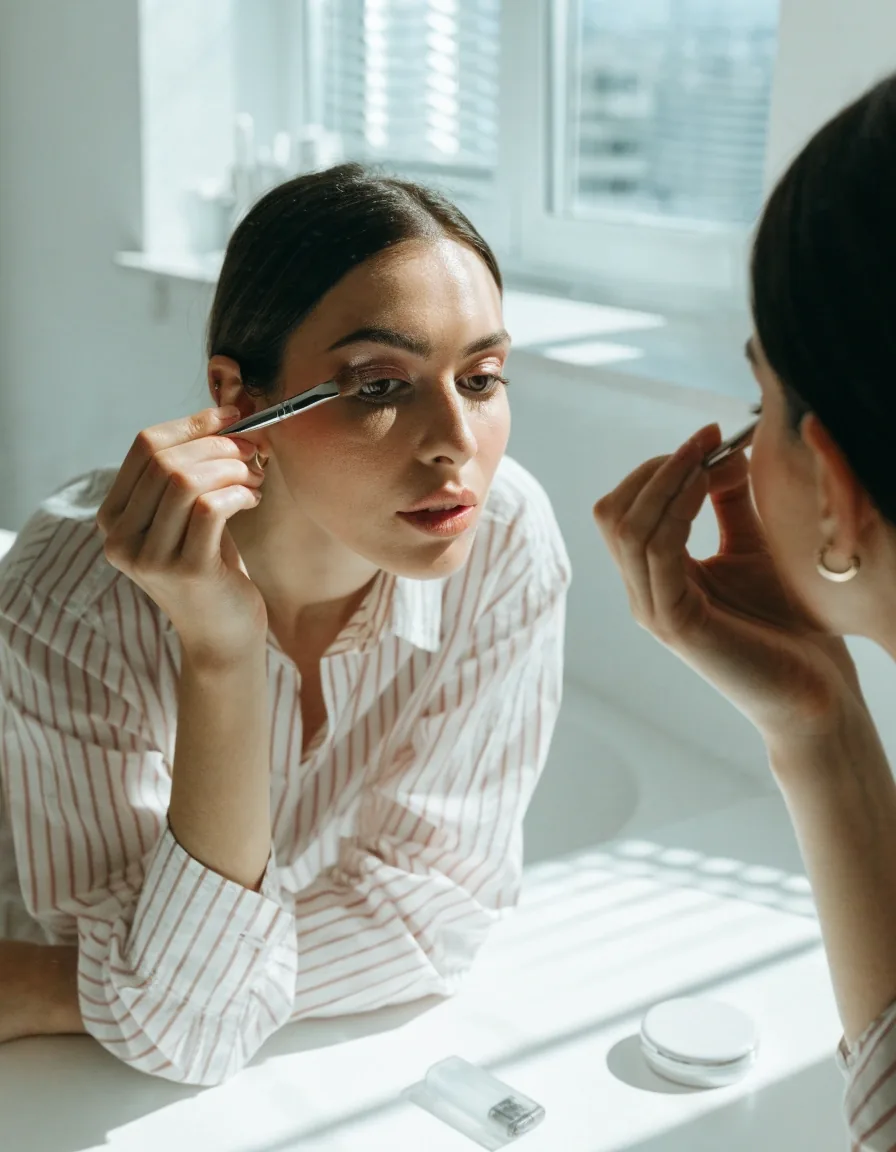 Woman applying eye makeup at a bright mirror with an open contact lens case on the counter